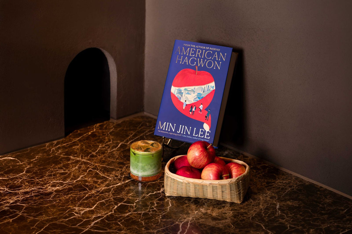 A book titled 'American Hagwon' by Min Jin Lee stands next to an apple cinnamon matcha and a basket of red apples The book cover is blue with a large red apple illustration.